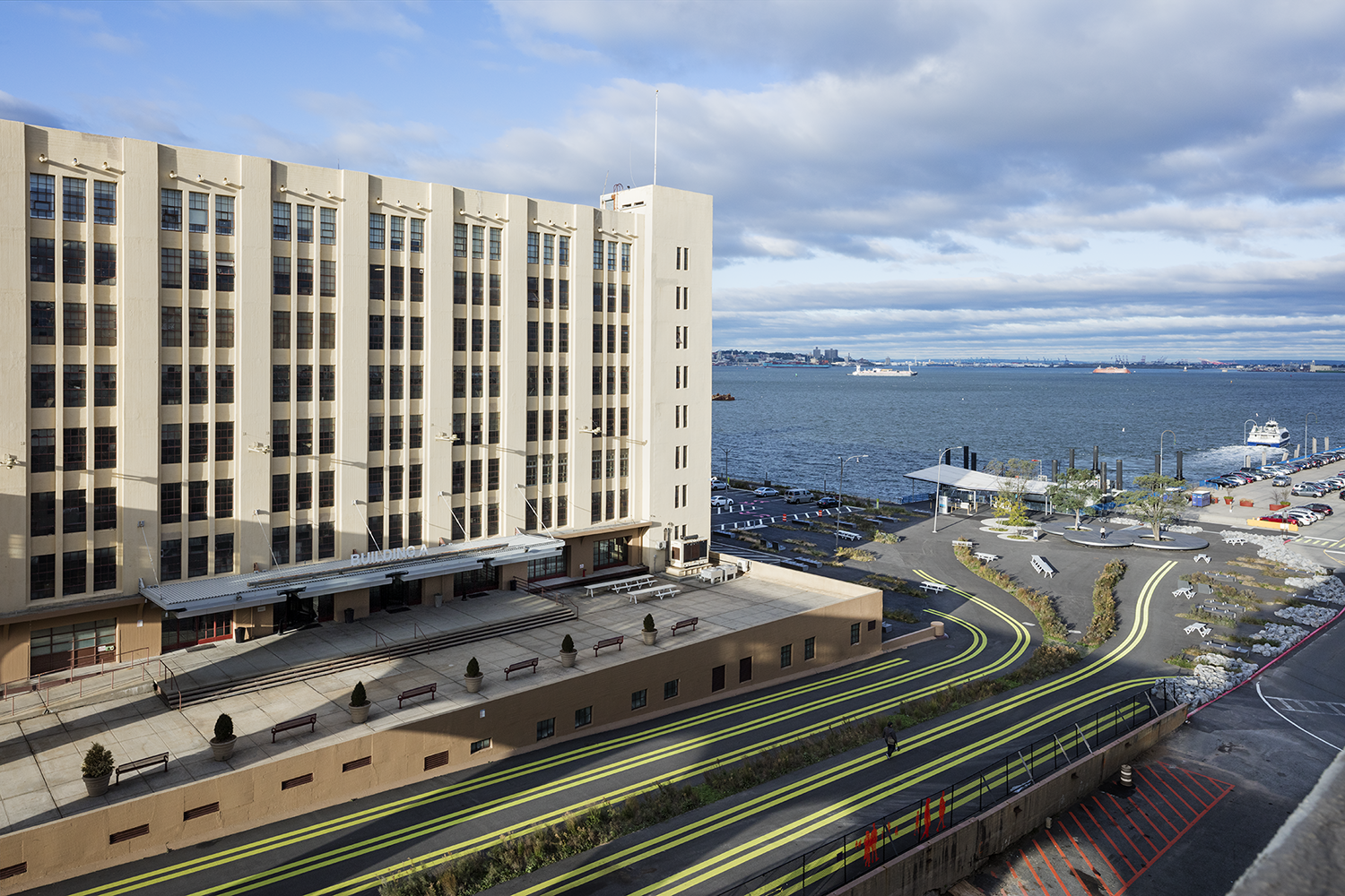 A waterfront view with a large, multi-story Brooklyn Army Terminal building on the left, to demonstrate the Climate Innovation Hub program. A road with yellow lines curves near the building. A parking area and dock are visible behind, with ships on the water and distant shoreline under a partly cloudy sky.