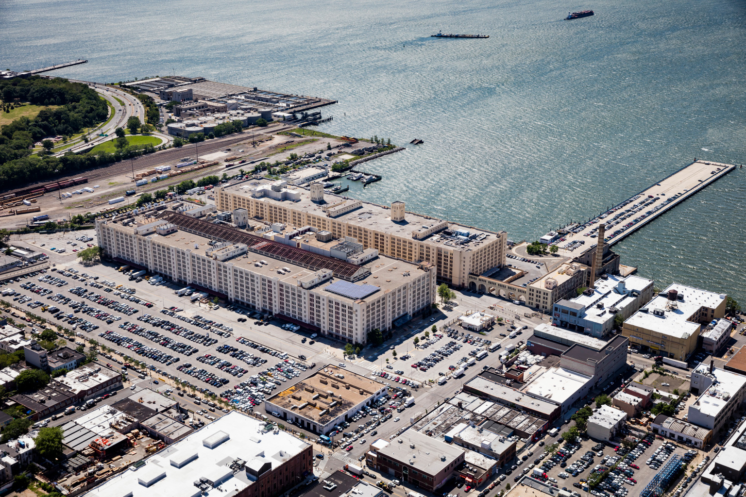 Aerial view of Brooklyn Army Terminal, a large waterfront warehouse complex with numerous parking lots filled with cars. The area is surrounded by industrial buildings, green spaces, and docks extending into the water.