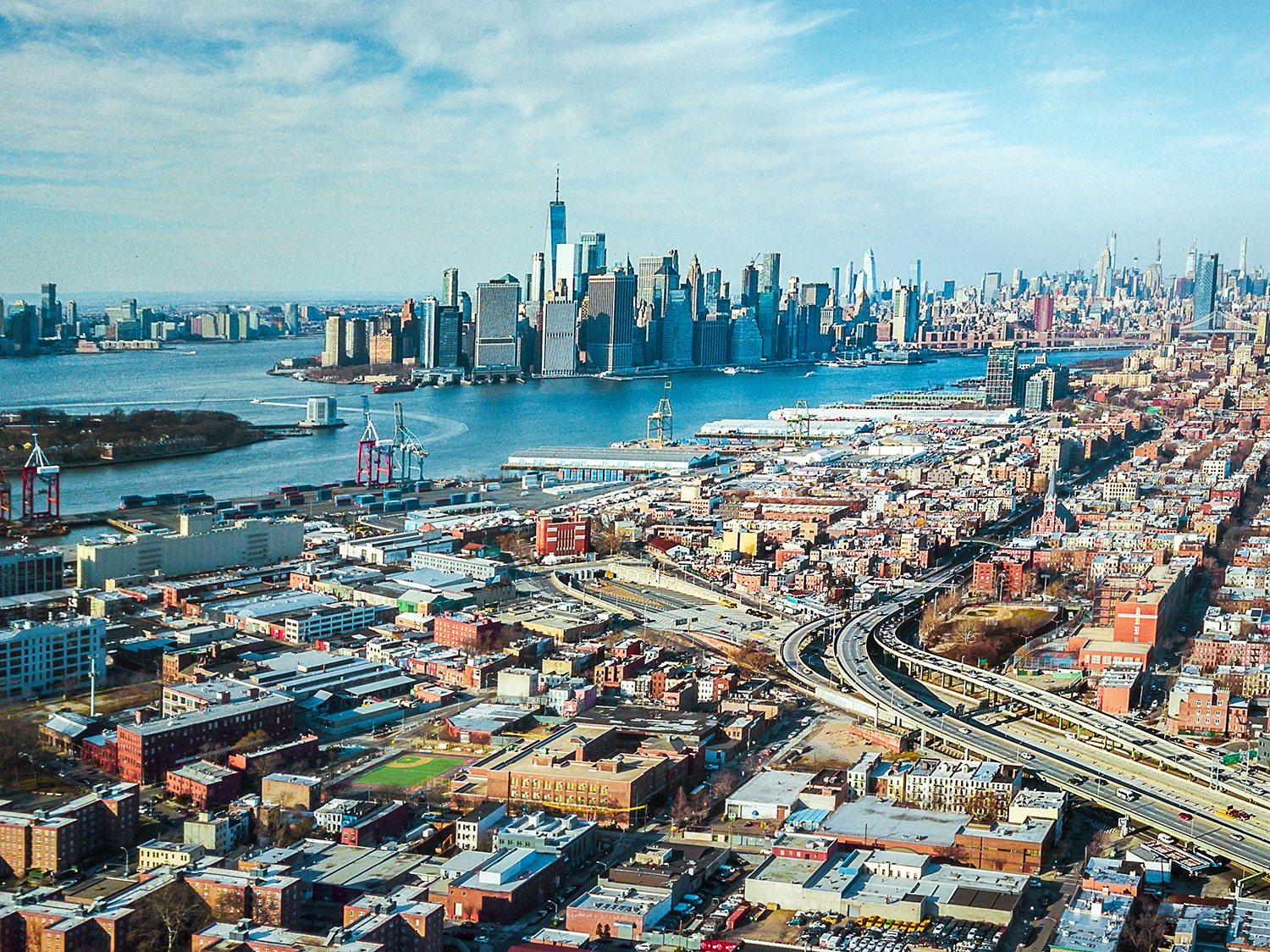 Aerial view of Red Hook Brooklyn featuring a mix of commercial, residential buildings, highways, and the New York Harbor with Manhattan skyscrapers in the background on a clear day.