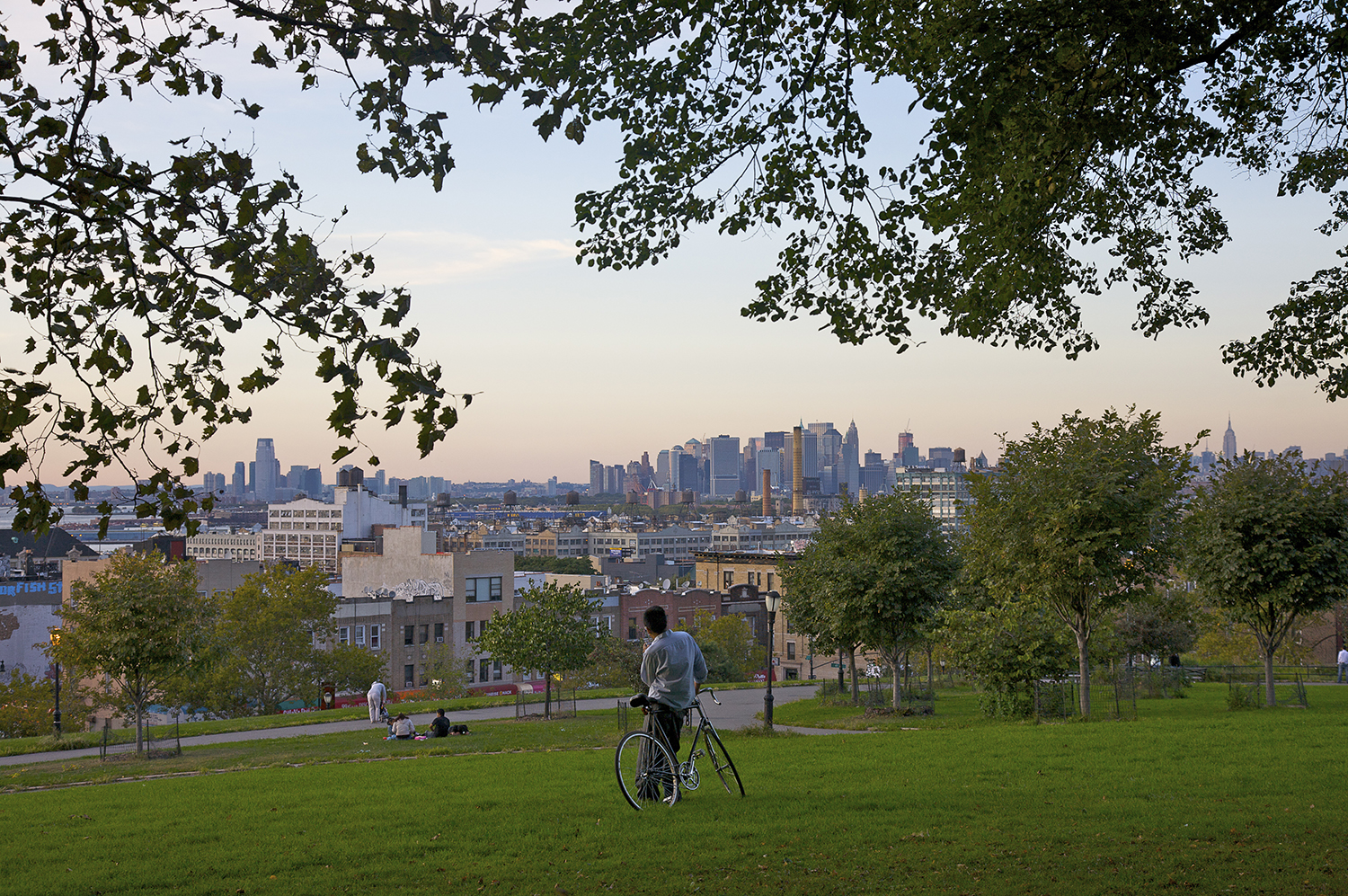 A person with a bicycle stands on a grassy hill in Sunset Park, overlooking a New York City skyline under a clear sky. Trees frame the scene.