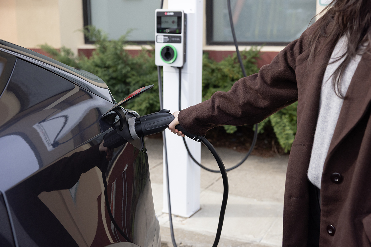 A person in a brown coat is charging an electric car at a BAT electric vehicle charging station. The charging cable is plugged into the car, and greenery is visible in the background.
