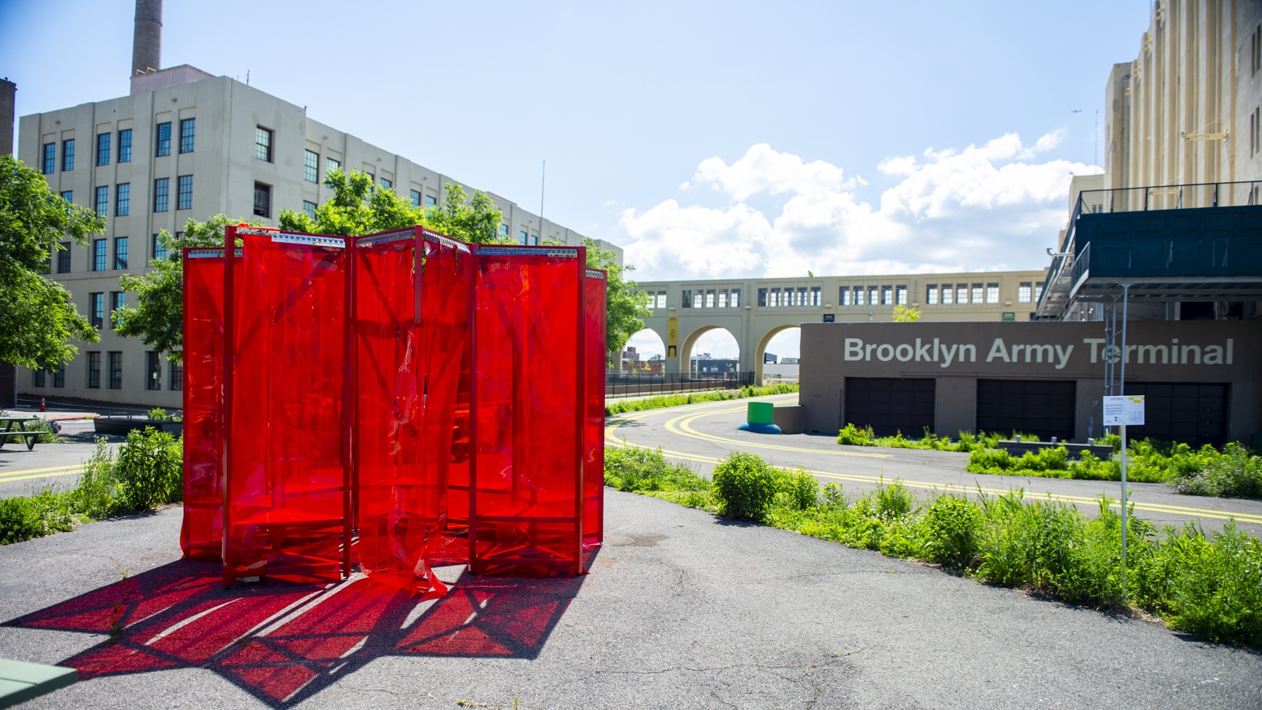 A large outdoor art installation made of translucent red panels stands in front of the Brooklyn Army Terminal, with a sunny sky, greenery, and industrial buildings in the background.
