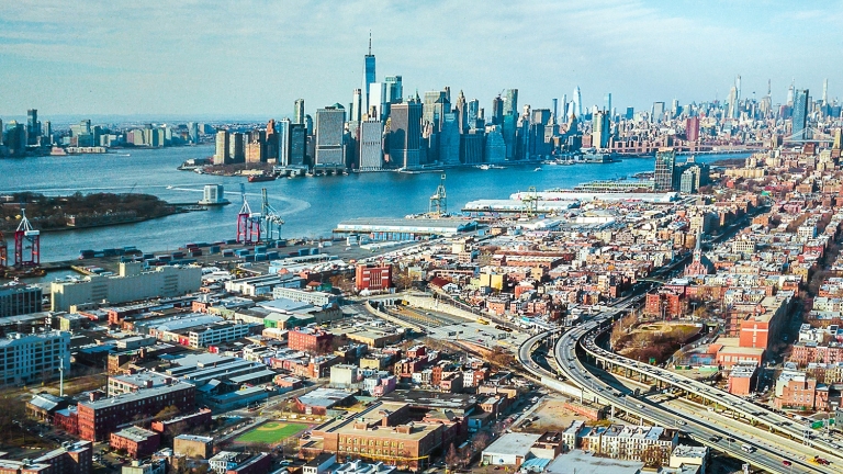 Aerial view of Red Hook Brooklyn featuring a mix of commercial, residential buildings, highways, and the New York Harbor with Manhattan skyscrapers in the background on a clear day.