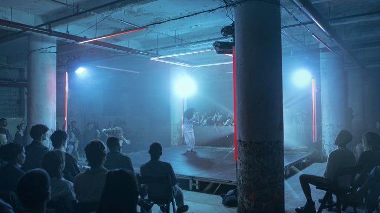  A dimly lit, atmospheric performance space with an audience seated around a small stage. A performer stands under blue lighting, surrounded by concrete columns and industrial architecture. Red neon lights accentuate the scene.