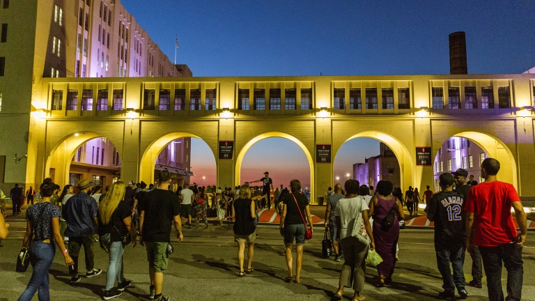 A crowd of people walk towards a lit building with large arches during dusk. The sky is a gradient of blue and orange, and the scene has an urban atmosphere.