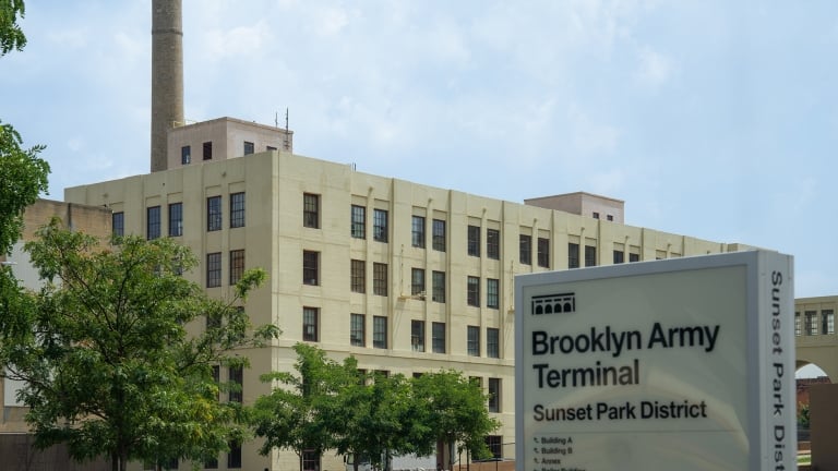 The image shows the Brooklyn Army Terminal building, a large beige industrial structure with a tall chimney. In the foreground, theres a sign reading Brooklyn Army Terminal, Sunset Park District and trees lining the entrance.