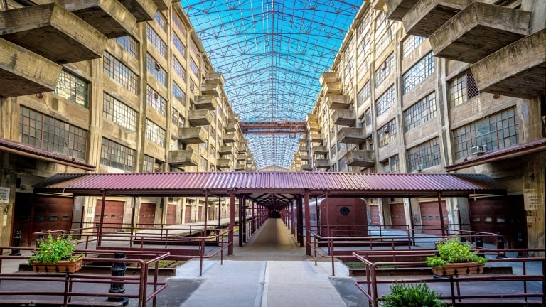 Brightly lit interior of a large industrial-style building with high glass ceilings, concrete beams, and metal railings. Rows of windows line the sides, and a central walkway is covered by a red roof. Small plants add a touch of green.