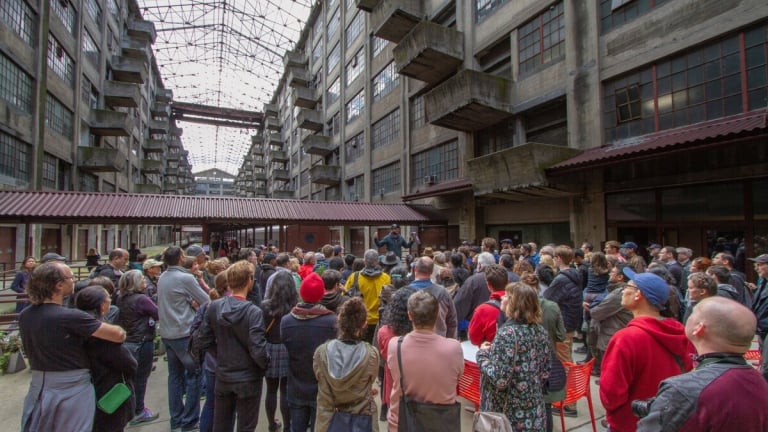  A large group of people gather in an industrial-style courtyard with high concrete walls and a glass ceiling. Some are seated on red chairs, while most stand and face a speaker. The atmosphere is lively and attentive.