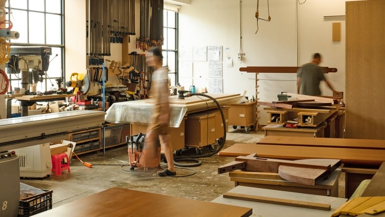 A woodworking workshop with two people working on projects. Various tools, wooden planks, and workbenches fill the space. Large windows allow natural light to illuminate the room. The scene is busy and organized.