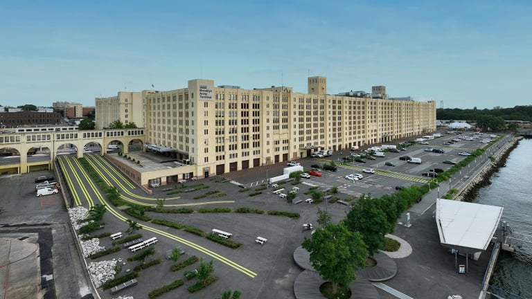 A large beige industrial building with vintage architecture sits by the waterfront. The surrounding area features a parking lot, landscaped greenery, walking paths, and a dock. The sky is clear and blue.