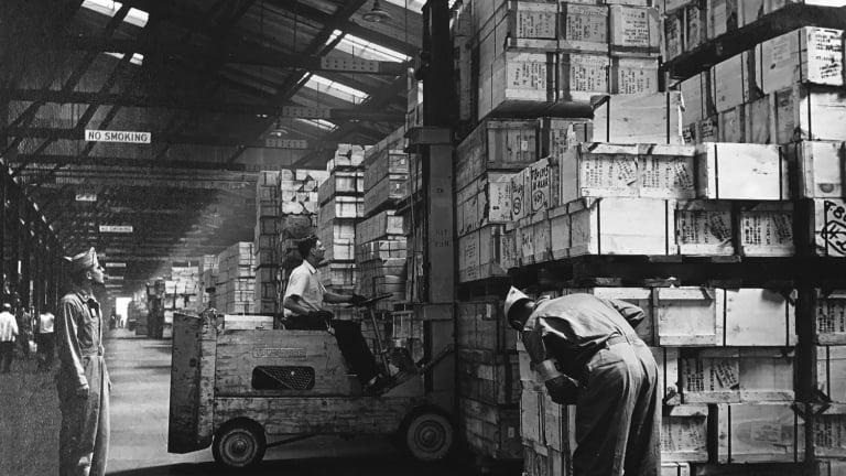 A black and white photo showing workers using a forklift to stack large crates in a warehouse. The warehouse has high ceilings and the crates are labeled with various markings. One person supervises while another inspects the crates.
