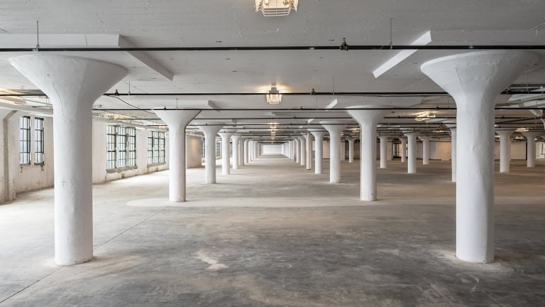 An empty, spacious industrial warehouse with white support columns and large windows along the walls. The concrete floor and ceiling are minimalist, and the space is well-lit by ceiling lights.