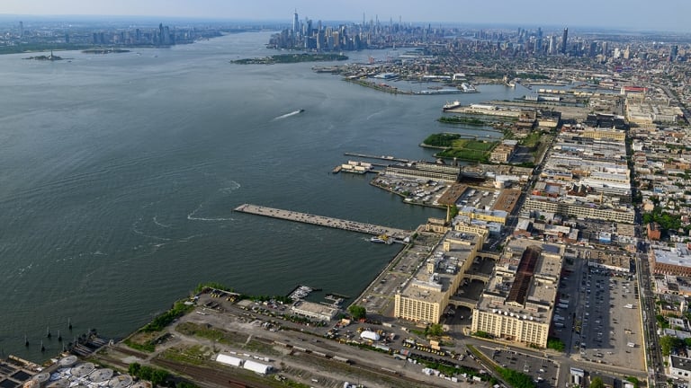 Aerial view of a cityscape featuring waterways and urban development. Skyscrapers are visible in the distance, with a large river dividing areas filled with industrial buildings and docks. Green spaces and roads are scattered throughout.