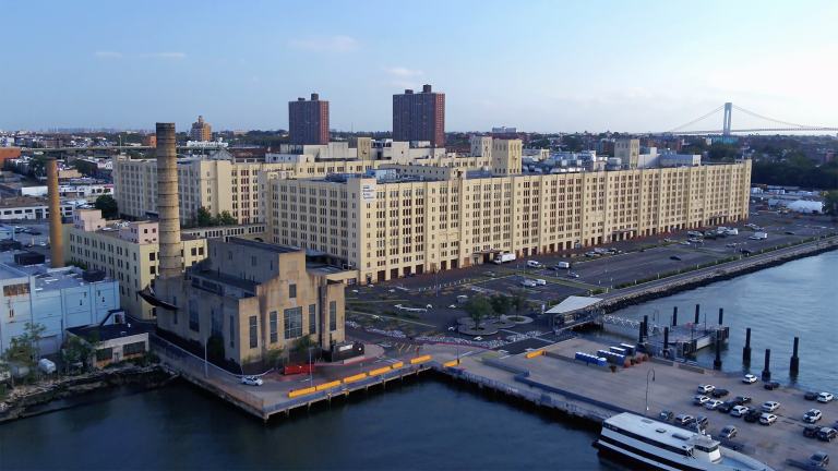 Aerial view of a waterfront industrial complex with a mix of modern and older buildings. A large factory-like structure is prominent, with residential high-rises in the background and a dock with vehicles in the foreground.