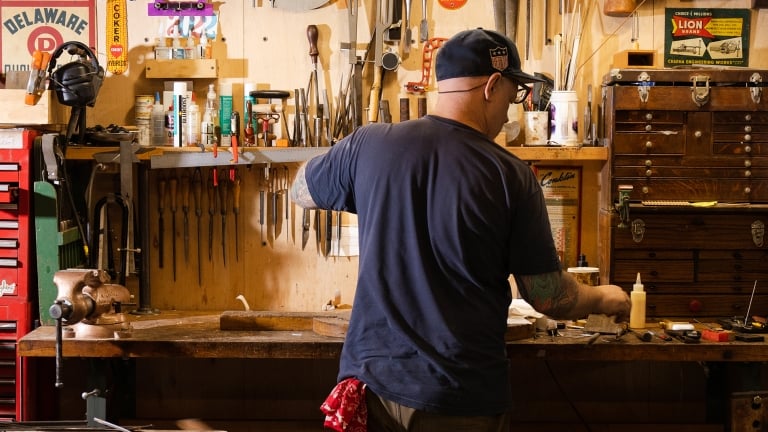 A person in a baseball cap works at a cluttered workshop bench with tools hanging on the wall. The space is filled with various equipment, including wrenches, pliers, and bottles. A red cloth hangs from their pocket.