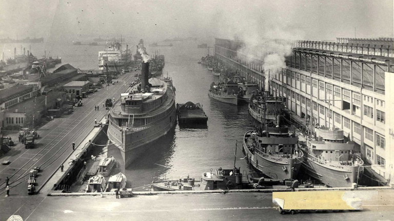 A vintage black-and-white photo of a busy harbor with multiple docked ships and tugboats. Smoke rises from one vessel, and warehouses line the right side. People and cargo are visible along the docks, with a foggy horizon in the background.