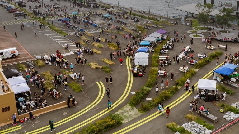 Aerial view of an outdoor food festival by the waterfront. Crowds of people gather around various food stalls and seating areas. The scene includes colorful tents and a view of the water with ships in the distance.