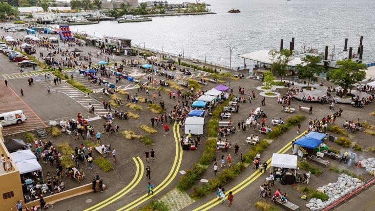 Aerial view of an outdoor food festival by the waterfront. Crowds of people gather around various food stalls and seating areas. The scene includes colorful tents and a view of the water with ships in the distance. Aerial view of an outdoor food festival by the waterfront. Crowds of people gather around various food stalls and seating areas. The scene includes colorful tents and a view of the water with ships in the distance.