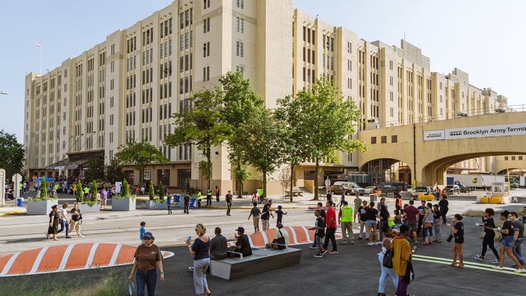 A bustling outdoor scene with people gathered near a large, historic building. Trees and artistic installations are in the foreground. The atmosphere is lively and sunny, with a mix of individuals socializing and walking in the area.