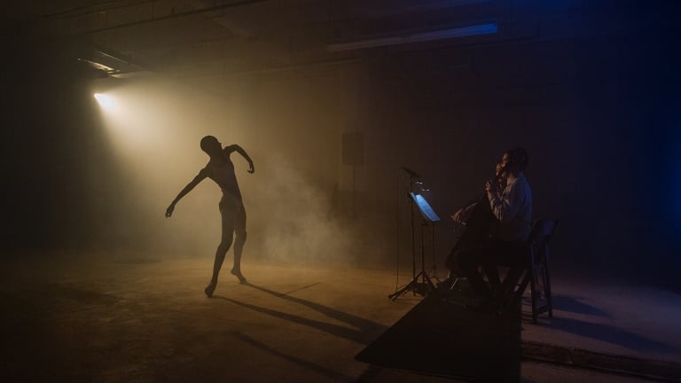 A dancer performs gracefully in a dimly lit room with a single spotlight, casting a dramatic shadow. In the background, a musician sits, playing a cello, adding to the moody atmosphere with smoke and ambient lighting.