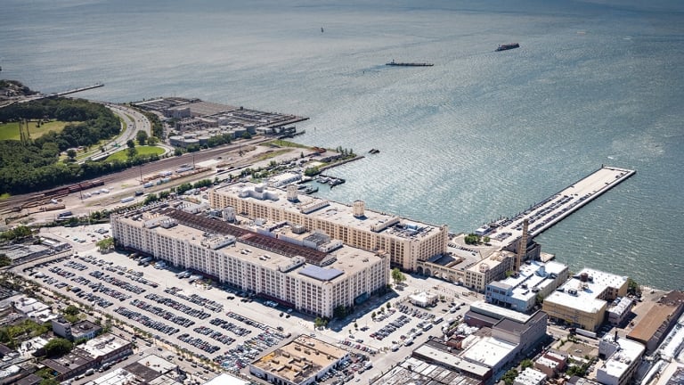 Aerial view of a large, multi-winged industrial building surrounded by parking lots. Located by the waterfront, with a pier extending into the water. Nearby roads and green spaces are visible, along with other buildings and infrastructure.