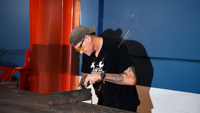 A person wearing a cap and protective glasses is using a tool to work on a metal piece, with orange sparks flying around. They have tattoos on their arms and are focused on the task.