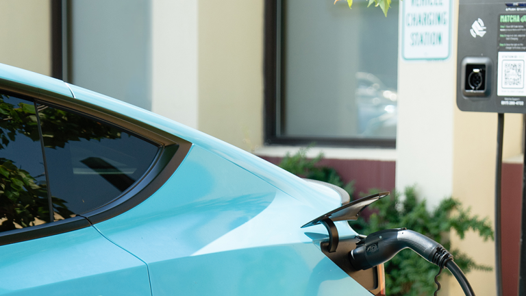 A light blue electric car is plugged into a charging station beneath a tree, with a green &quot;Matcha Electric Vehicle Charging Station&quot; sign visible in the background, outside of the Brooklyn Army Terminal.