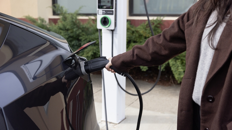A person in a brown coat is charging an electric car at a BAT electric vehicle charging station. The charging cable is plugged into the car, and greenery is visible in the background. A person in a brown coat is charging an electric car at a BAT electric vehicle charging station. The charging cable is plugged into the car, and greenery is visible in the background.