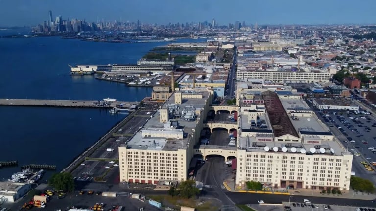 Aerial view of a waterfront industrial complex with large white buildings, parking lots, and a pier, with a city skyline and water in the background under a clear blue sky.