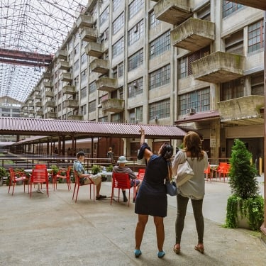 People gather in an industrial courtyard with red chairs and tables. Two women stand pointing upwards, while two men sit conversing. The space is surrounded by multi-story building sections with balconies and an open roof. People gather in an industrial courtyard with red chairs and tables. Two women stand pointing upwards, while two men sit conversing. The space is surrounded by multi-story building sections with balconies and an open roof.