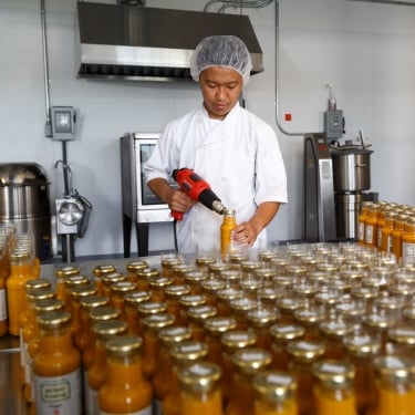 A person in a hairnet and white coat uses a drill-like tool to seal bottles filled with an orange liquid in a factory setting. Numerous sealed bottles are lined up on a metal table. Industrial kitchen equipment is visible in the background.