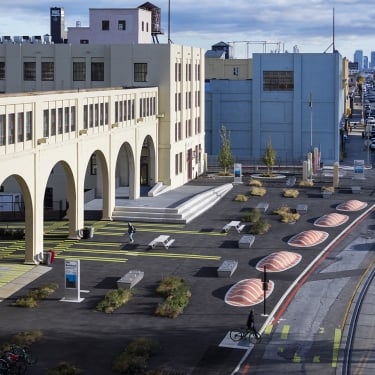 Urban park with modern design features between large industrial buildings. Includes pathways, benches, landscaped greenery, and striped patterns on the ground. A cyclist rides along the street, with a city skyline visible in the distance.