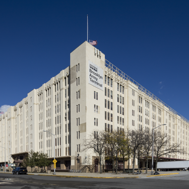A large, white industrial building with multiple stories, identified as the Brooklyn Army Terminal. The structure has a slightly angular design, with an American flag on top. The sky is clear and blue. Trees and vehicles are visible around the building.