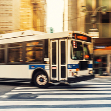 A city bus in motion travels through an urban area with tall buildings in the background. The bus is white with a blue stripe and is crossing a pedestrian crosswalk on a city street. The photo has a blurred effect. A city bus in motion travels through an urban area with tall buildings in the background. The bus is white with a blue stripe and is crossing a pedestrian crosswalk on a city street. The photo has a blurred effect.