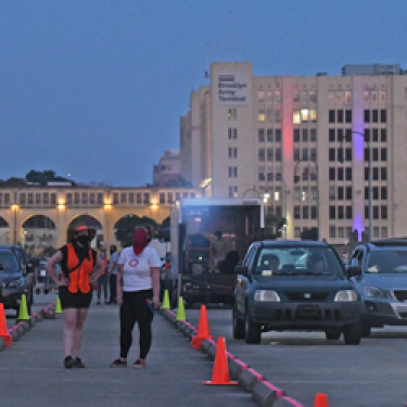 People walking and cars lined up between traffic cones on a street at dusk. In the background, a large building with Brooklyn Army Terminal on it is illuminated with colorful lights.