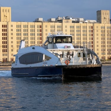 A blue and white ferry travels on a body of water with a background of tall, beige buildings under a clear sky. A blue and white ferry travels on a body of water with a background of tall, beige buildings under a clear sky.
