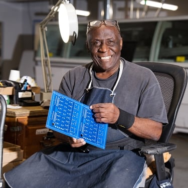  A man sitting in a workshop smiles while holding a blue circuit board. He wears a black apron, a wristband, and has glasses on his head. The background features tools and industrial equipment.