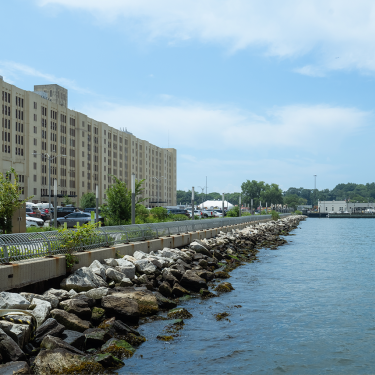 A long building lines a waterfront path with parked cars and rocky shore. The sky is partly cloudy, and calm water stretches to the right. Trees and small buildings are visible in the distance. A long building lines a waterfront path with parked cars and rocky shore. The sky is partly cloudy, and calm water stretches to the right. Trees and small buildings are visible in the distance.