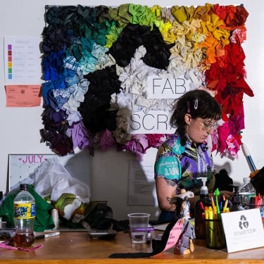 A person is working at a desk covered with various items, including drinks and papers. Behind them is a colorful, textured fabric art piece on the wall, labeled Fab Scrap. The scene is brightly lit with a mix of colors and shadows.