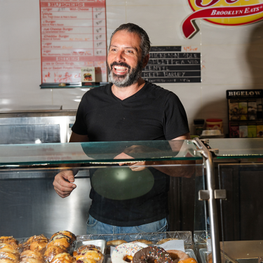 A man with a beard and a black T-shirt stands behind a glass display filled with pastries. Hes smiling and standing in a bakery named Pete&#039;s Brooklyn Eats, as indicated by the sign on the wall behind him.