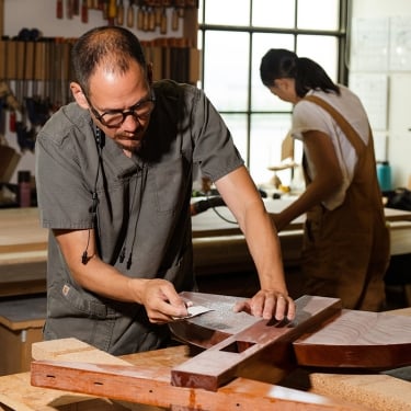 A man wearing glasses works on a wooden project in a workshop, using a hand tool. In the background, another person is focused on their task. The room is well-lit with natural light coming through a large window.