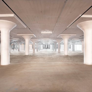 A large, empty industrial warehouse space with concrete floors and ceiling. White columns evenly spaced throughout the room, with multiple windows on the back wall allowing natural light to fill the space.