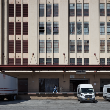 A large industrial building with a loading dock features two white vans parked in front. A man sits on the dock, and a truck trailer is visible on the left. The building has numerous windows and a beige exterior. A large industrial building with a loading dock features two white vans parked in front. A man sits on the dock, and a truck trailer is visible on the left. The building has numerous windows and a beige exterior.
