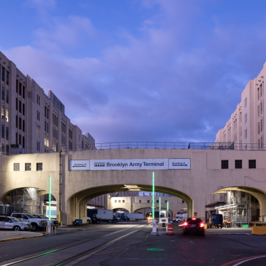 View of the Brooklyn Army Terminal at dusk, featuring large industrial buildings with arched entrances. The road leads through the structure, with some parked vehicles and visible construction work under the lit sky.