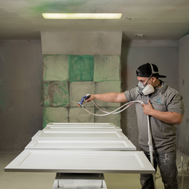 A person wearing a cap and a protective mask is spray painting cabinet doors in a workshop. The room has a ventilation system with filters on the wall and overhead lights illuminating the workspace.