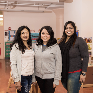 Three women stand smiling inside a classroom, featuring colorful educational materials and childrens furniture. The room has a warm, welcoming atmosphere with educational posters on the walls.