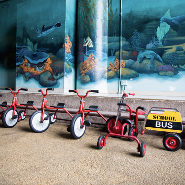 Five red tricycles are lined up under an ocean-themed mural, along with a tricycle attached to a yellow cart labeled School Bus.