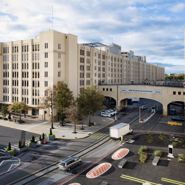 A large Art Deco-style building with multiple stories and tall, narrow windows. A roadway passes beneath an arched bridge that connects the building sections. Trees and parked cars are visible, and the sky is partly cloudy.