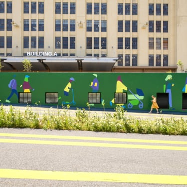 A green mural on a wall depicts colorful silhouettes of people walking and pushing strollers. A person in an orange outfit walks past in front of the mural. Behind the wall is a beige industrial building labeled “Building A.”.