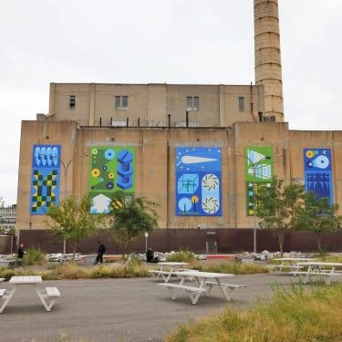 A large industrial building with colorful abstract murals on its exterior walls, depicting geometric shapes and patterns. There are trees, grass, and several white picnic tables in the foreground.